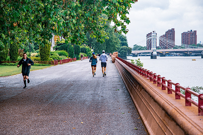 The image depicts a scenic riverside path where several people are engaged in running or walking. The path is lined with trees on one side and a railing on the other, with a bridge and buildings visible in the background.