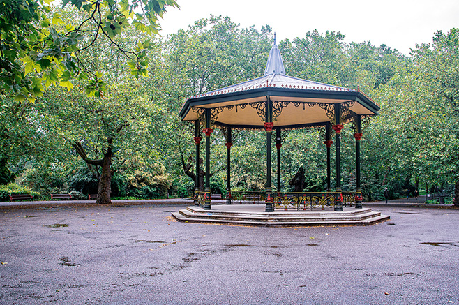 The image shows an ornate gazebo situated in a park. The gazebo is octagonal with a decorative roof and railings, and it is surrounded by trees and a paved area. The scene appears to be on a rainy day, as the ground is wet and there are puddles visible.