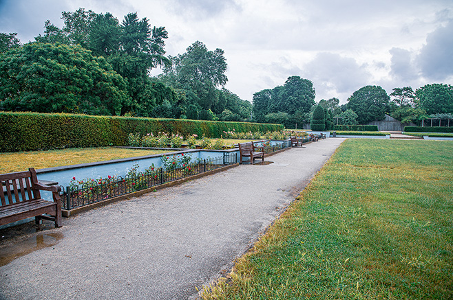 The image depicts a serene park scene with a paved walkway bordered by a well-maintained lawn on one side and a flower bed with a small water channel on the other. The park features several benches along the path, offering spots for rest and contemplation. Lush greenery, including trees and hedges, surrounds the area, creating a tranquil and inviting atmosphere.