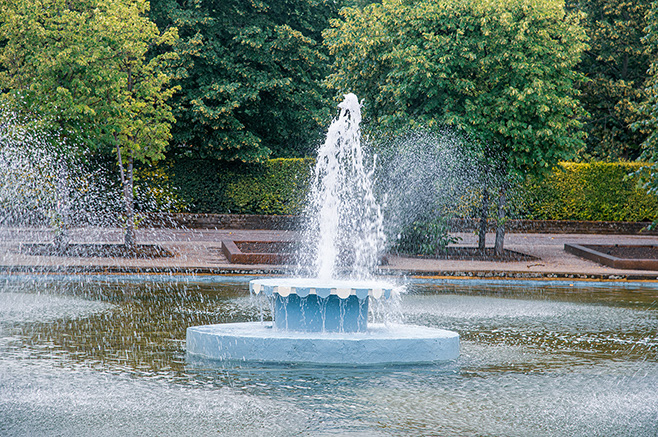 The image depicts a serene park scene with a prominent water fountain in the center of a pond. The fountain is surrounded by lush greenery and trees, creating a tranquil and refreshing atmosphere.