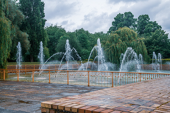 The image depicts a serene park scene with multiple water fountains spraying water in a semi-circular arrangement. The fountains are surrounded by a metal railing and a brick-paved walkway. In the background, there are lush green trees under a cloudy sky, creating a tranquil and inviting atmosphere.
