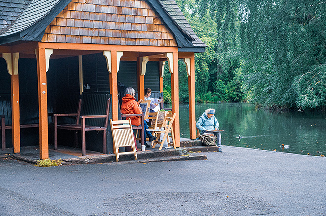 The image shows a serene lakeside scene with a wooden pavilion. Three people are seated near the water, engaged in activities such as fishing and relaxing. The pavilion is situated on a paved area adjacent to the lake, surrounded by lush greenery.