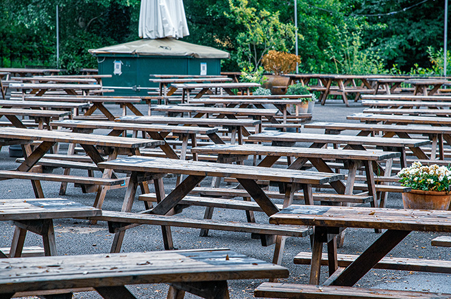 The image shows an outdoor seating area with numerous wooden picnic tables arranged in rows. The area is surrounded by greenery, and there is a small structure or hut in the background. Some tables have potted plants on them, adding a touch of decoration. The ground is covered with gravel, and the overall setting appears to be a park or a similar public space.