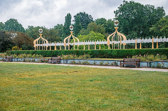 The image depicts a well-maintained garden area with ornate, decorative structures featuring domed tops and intricate designs. The structures are arranged in a row, separated by a hedge and a pathway. Benches are placed along the pathway, inviting visitors to sit and enjoy the view. The garden is lush with greenery, and trees are visible in the background, adding to the serene atmosphere.