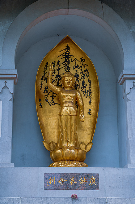 The image shows a golden statue of a Buddha figure placed in a niche with an arch. The statue is adorned with intricate details and is surrounded by Chinese characters inscribed on the backdrop. The statue stands on a lotus base, symbolizing purity and enlightenment. Below the statue, there is a plaque with additional Chinese characters, and a single flower is placed at the base, likely as an offering.
