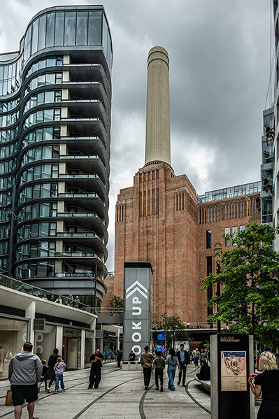 The image depicts an urban scene with a mix of modern and historical architecture. A tall, cylindrical chimney stands prominently in the background, likely part of an old industrial building. Adjacent to it is a modern building with curved balconies. The area appears to be a public space with people walking around, and there is signage indicating a 'LOOK UP' campaign, encouraging people to appreciate their surroundings.