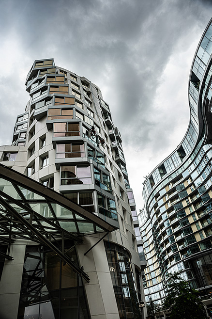 The image depicts modern, uniquely designed buildings with a mix of geometric and curved shapes. The architecture features a variety of materials, including glass and metal, and the buildings are closely packed together. The sky above is overcast, adding a dramatic effect to the scene. The overall atmosphere is urban and contemporary.