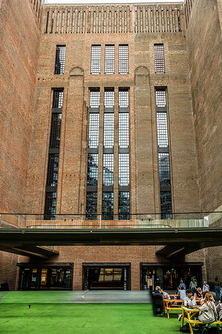 The image depicts a large, historic building with tall, arched windows and a brick facade. In the courtyard in front of the building, there is a grassy area and a group of people seated at tables, possibly having a meal or meeting.