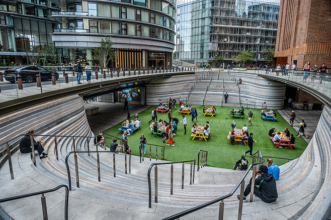 The image depicts a modern urban setting with a unique architectural design. In the center, there is a sunken area with a grassy lawn where people are sitting and socializing. Surrounding this area are curved, tiered stone steps where more individuals are seated. The scene is framed by contemporary buildings with large glass windows, indicating a bustling, possibly commercial, environment. The overall atmosphere appears lively and communal, with people enjoying the outdoor space.