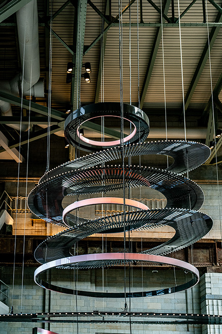 The image depicts a modern, multi-tiered, circular chandelier suspended from the ceiling of a large, industrial-style room. The chandelier consists of several concentric rings, each adorned with small lights, and is hung by multiple cables. The room features exposed structural elements such as beams and ducts, contributing to an industrial aesthetic.