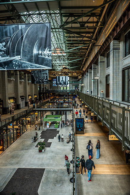 The image depicts a bustling indoor space, likely a train station or a large public transportation hub. The architecture features high ceilings with exposed metal beams and large windows allowing natural light to flood the area. There are multiple levels with walkways and balconies, and people are seen walking, standing, and engaging in various activities. The space includes various shops and kiosks, and there is a large digital display hanging from the ceiling. The overall atmosphere is busy yet organized, with a modern and industrial aesthetic.