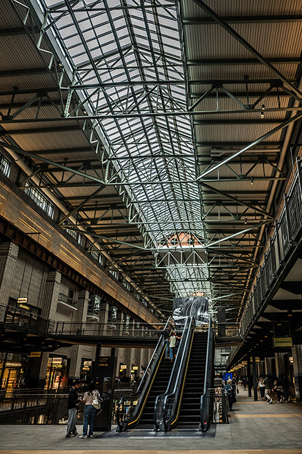 The image depicts a large, modern indoor space with a high, glass-paneled ceiling supported by a metal framework. The area features escalators connecting different levels, with people using them and walking around. The space appears to be a shopping mall or a similar commercial area, with various stores and seating areas visible on the lower level.