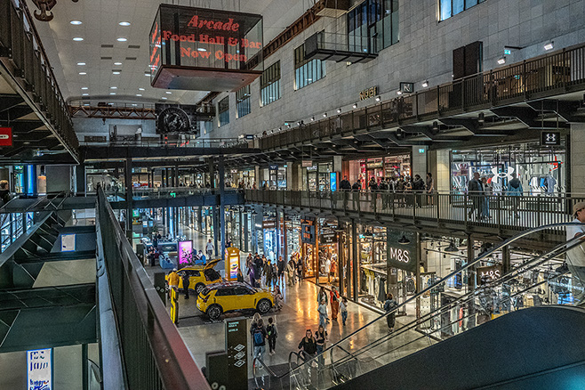 The image depicts a bustling multi-level shopping mall with various stores and a prominent sign advertising an arcade food hall on the upper level. The mall features modern architecture with glass railings, escalators, and a variety of retail outlets, including a visible M&S store. Shoppers can be seen walking around, and there are bright lights and colorful displays throughout the space.