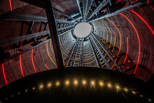 The image depicts the interior of a large, cylindrical structure with a complex network of stairs and walkways. The structure is illuminated with red and yellow lights, creating a striking visual effect. The perspective is from the bottom, looking upwards towards the top of the cylinder, which has a circular opening. The design is industrial and architectural, suggesting it could be part of a large public or commercial building.