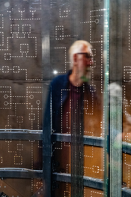 he image shows a blurred view of a person wearing a white hat and a dark jacket, seen through a window with a grid pattern and condensation. The person appears to be indoors, possibly in a public transportation setting like a bus or train.