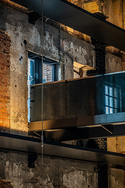 The image depicts an industrial or warehouse setting with exposed brick walls, concrete surfaces, and visible structural elements such as beams and pipes. There are windows with blue frames, and the lighting suggests it is either early morning or late afternoon, casting a warm glow on the rough textures of the walls and beams.