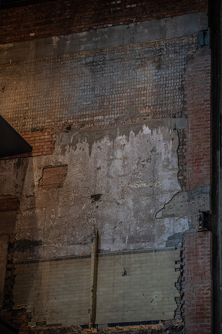 The image shows a dilapidated wall with exposed brick and plaster, indicating significant wear and tear. The wall has a large section where the plaster has fallen off, revealing the brickwork underneath. The area appears to be in a state of disrepair, with debris and remnants of the fallen plaster scattered on the floor.