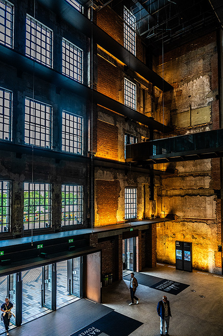 The image depicts the interior of a large, industrial-style building with exposed brick walls and large windows. The space is illuminated with warm lighting, highlighting the architectural details. There are a few people walking around, and the area appears to be part of a public or commercial space, possibly a museum or gallery.