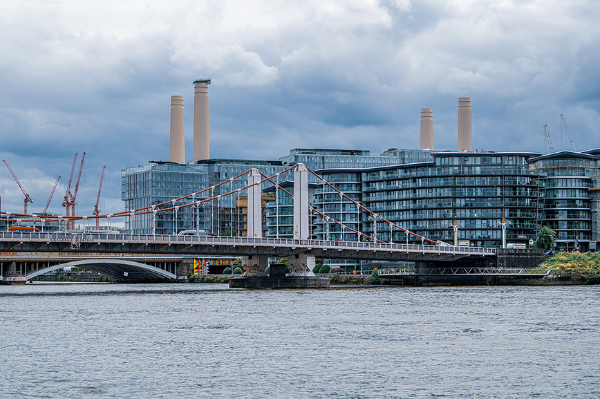 The image depicts a modern urban scene featuring a large, contemporary building complex with multiple towers and smokestacks. In the foreground, there is a bridge spanning a body of water, with cranes visible, indicating ongoing construction or development in the area. The sky is overcast, suggesting a cloudy day.