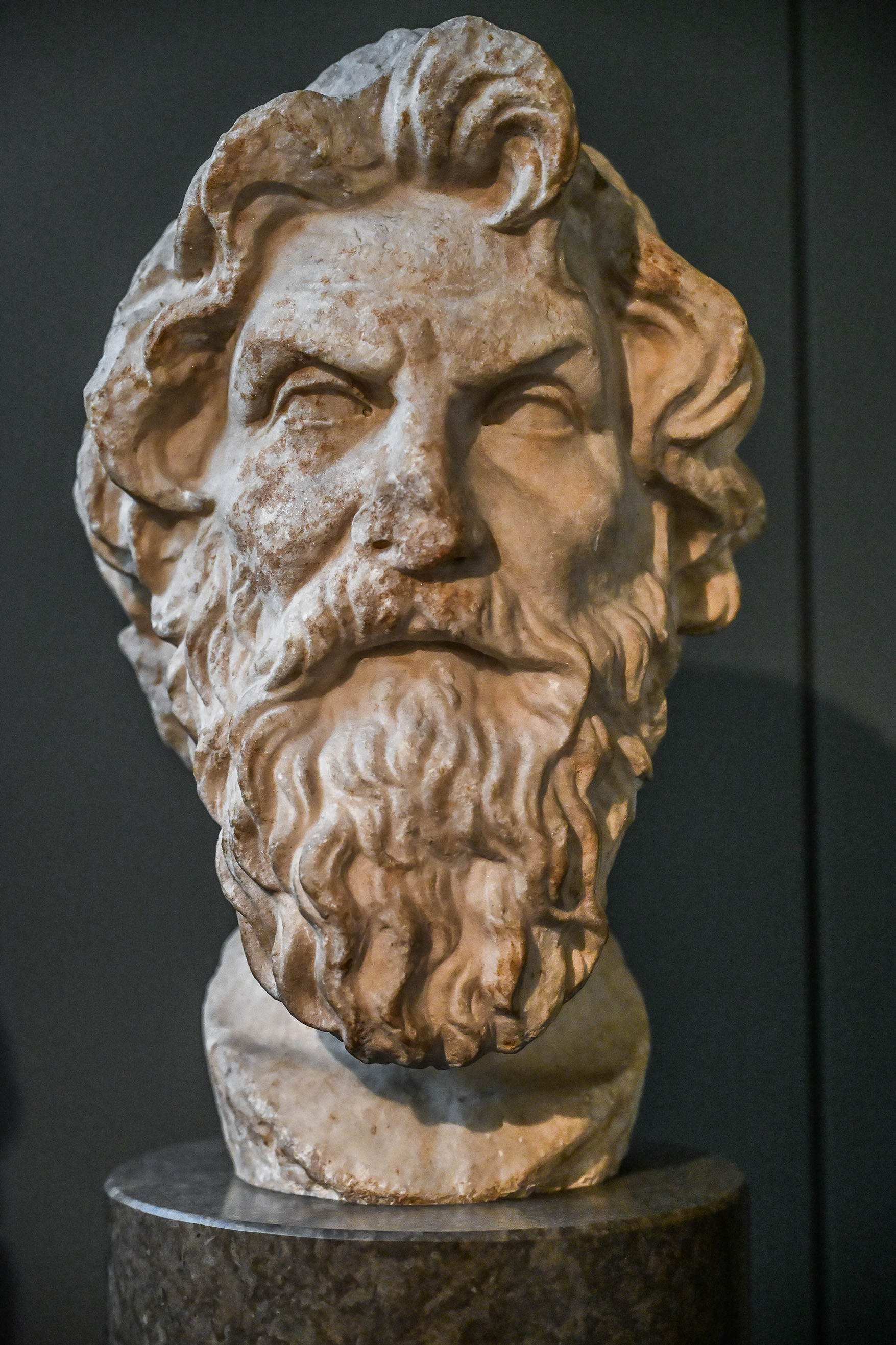 The image depicts a stone bust of an elderly man with a long, curly beard and wavy hair. The sculpture appears to be an ancient or classical artwork, characterized by detailed facial features and a serene expression. The bust is mounted on a cylindrical pedestal, and the background is dark, which accentuates the details of the sculpture.