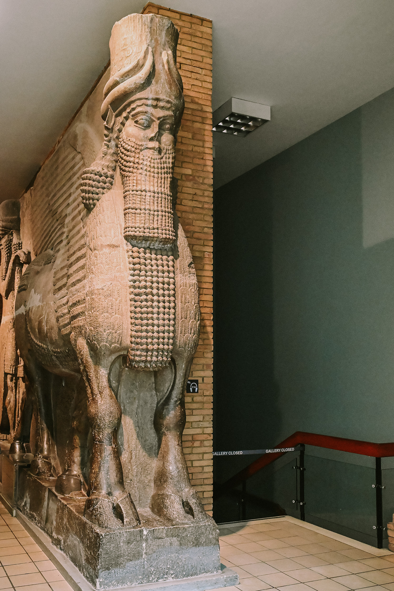 The image shows a large, intricately carved stone statue of a winged figure with a human head and a bull's body, known as a lamassu. This statue is displayed in a museum setting, with a brick wall behind it and a modern gallery environment around it. The lamassu has detailed features, including a beard, headdress, and intricate clothing. The gallery appears to be closed, as indicated by a sign on a glass door.
