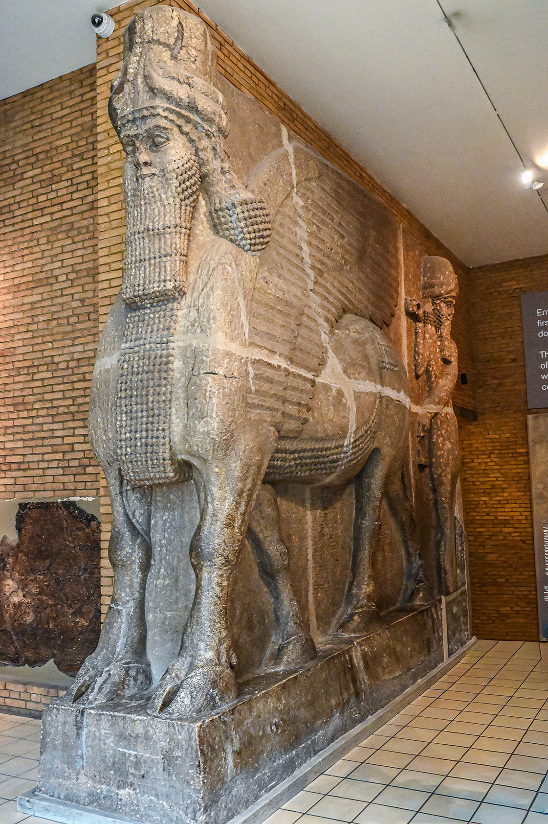 The image depicts an ancient bas-relief sculpture, likely of Assyrian origin, showcasing a winged figure with detailed features and intricate craftsmanship. The sculpture is displayed in a museum setting with brick walls and tiled flooring. The figure appears to be part of a larger relief panel, with other figures visible in the background. The craftsmanship and style suggest it is a significant historical artifact.