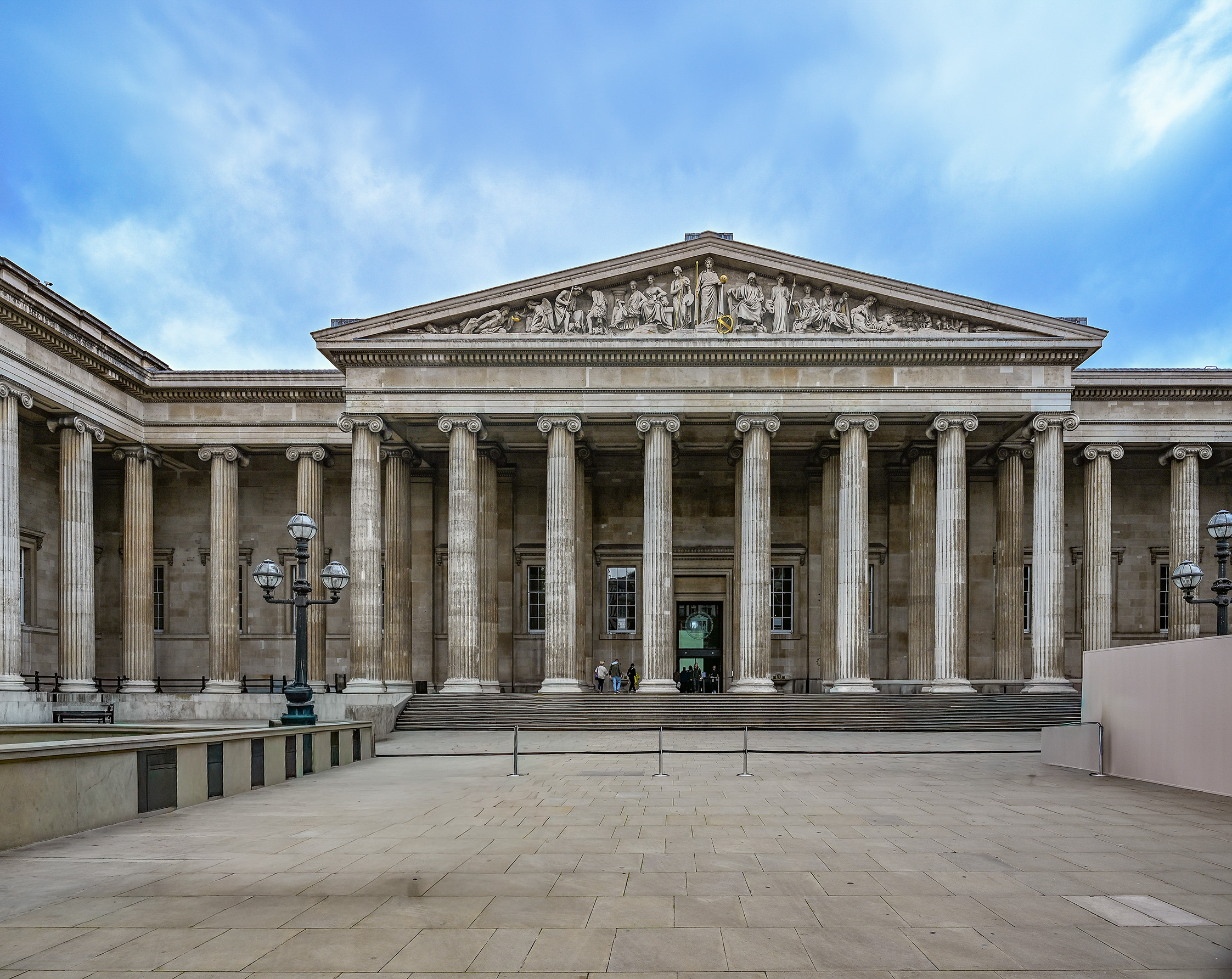 The image depicts the front facade of a grand neoclassical building, characterized by its large, imposing columns and intricate pediment. The structure features a series of tall, fluted columns with ornate capitals, supporting a triangular pediment adorned with detailed sculptures. The entrance is accessed by a set of wide steps, leading up to large doors. The sky above is partly cloudy, and the area in front of the building is an open, paved plaza with a few people visible near the entrance.