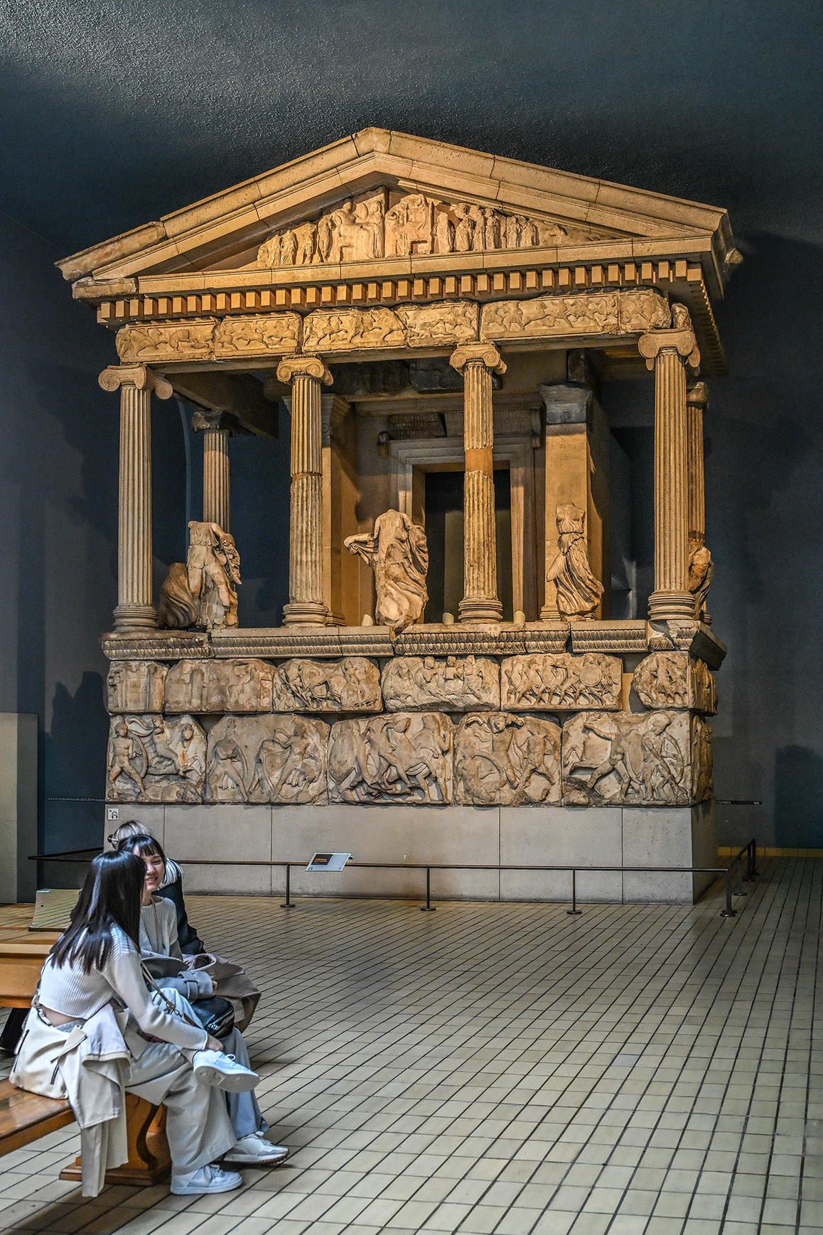 The image shows two individuals sitting and observing an ancient Greek temple-like structure with intricate carvings and statues. The structure features classical architectural elements such as columns and detailed friezes, likely depicting mythological scenes. The setting appears to be an indoor museum or exhibition space, with the individuals seemingly engaged in a discussion or admiring the historical artifact.