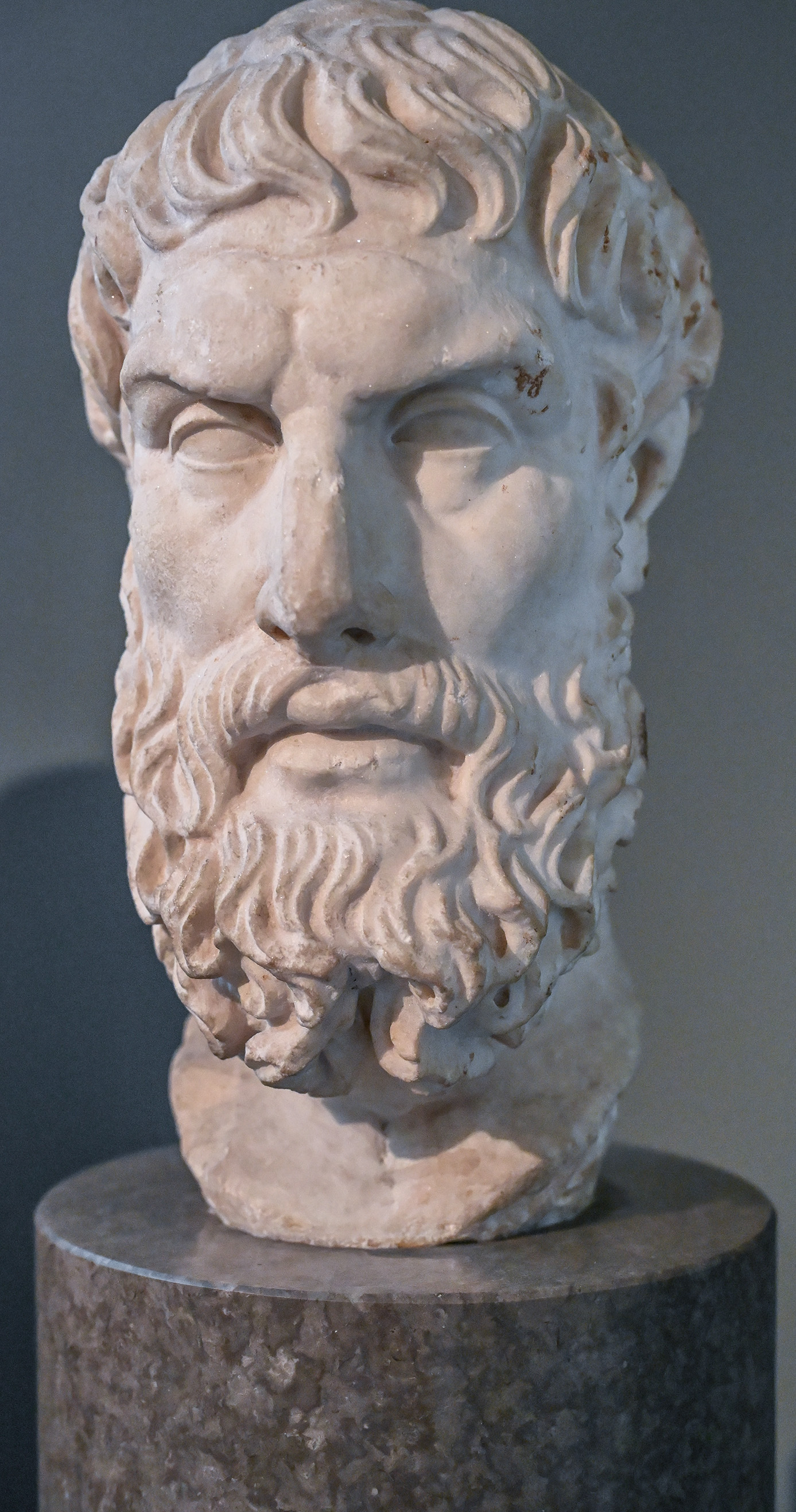 The image shows a marble bust of an ancient figure, likely from classical antiquity. The bust depicts a bearded man with curly hair, characterized by detailed craftsmanship. The eyes appear to be hollow, a common feature in ancient sculptures. The bust is mounted on a cylindrical pedestal, suggesting it is part of a museum or historical collection.