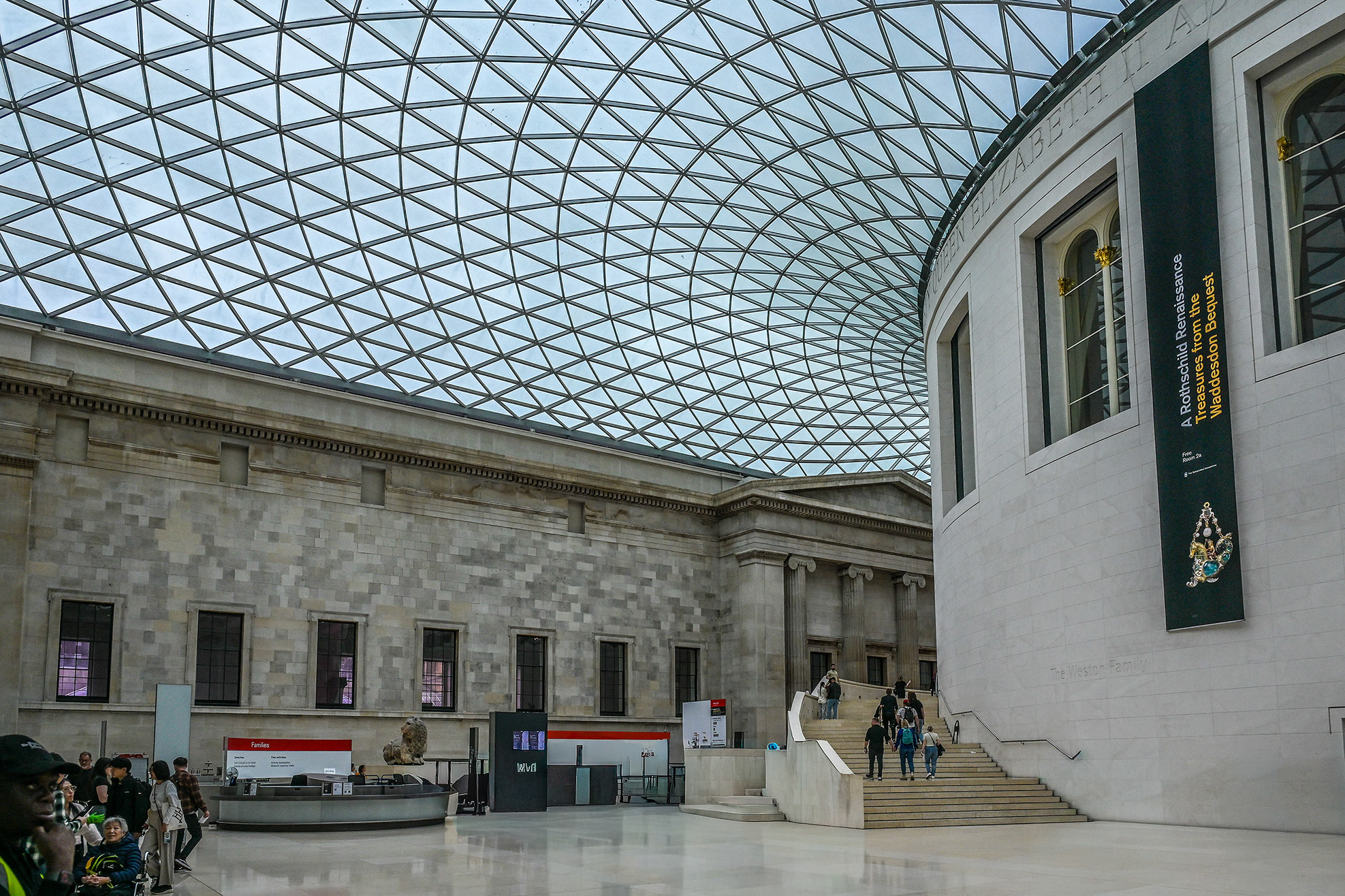 The image depicts the interior of a large, modern building with a distinctive glass roof composed of a geometric grid pattern. The space features classical architectural elements such as stone walls, columns, and large windows. There are several people in the area, some walking up a set of stairs and others gathered around a central information desk. The overall atmosphere is bright and spacious, indicative of a public or cultural institution, likely a museum or gallery.
