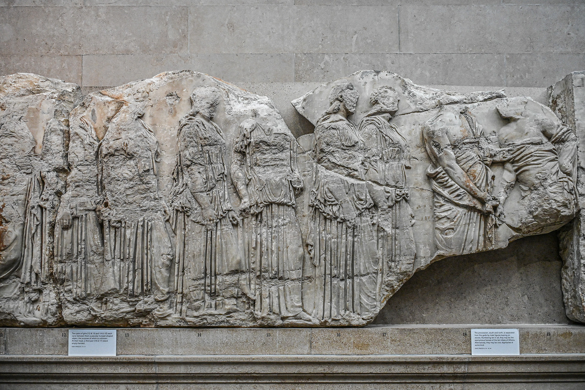 The image depicts an ancient Greek frieze, a sculpted relief originally part of a larger architectural structure. It features a procession of robed figures, likely from classical antiquity, showcasing intricate detailing of their garments and postures. The figures appear to be in a ceremonial or religious context, reflecting the artistic style and cultural themes of ancient Greek art.