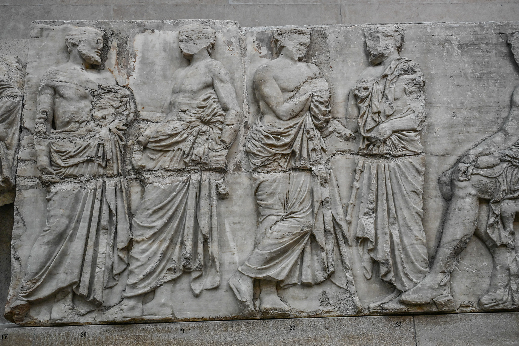 The image depicts an ancient Greek relief sculpture featuring a group of figures in flowing robes, likely goddesses or important women, standing in a row. The sculpture showcases intricate details of drapery and form, characteristic of classical Greek art. The figures are partially eroded, indicating its age and historical significance. This type of artwork is often found in museums or historical sites dedicated to ancient Greek culture.