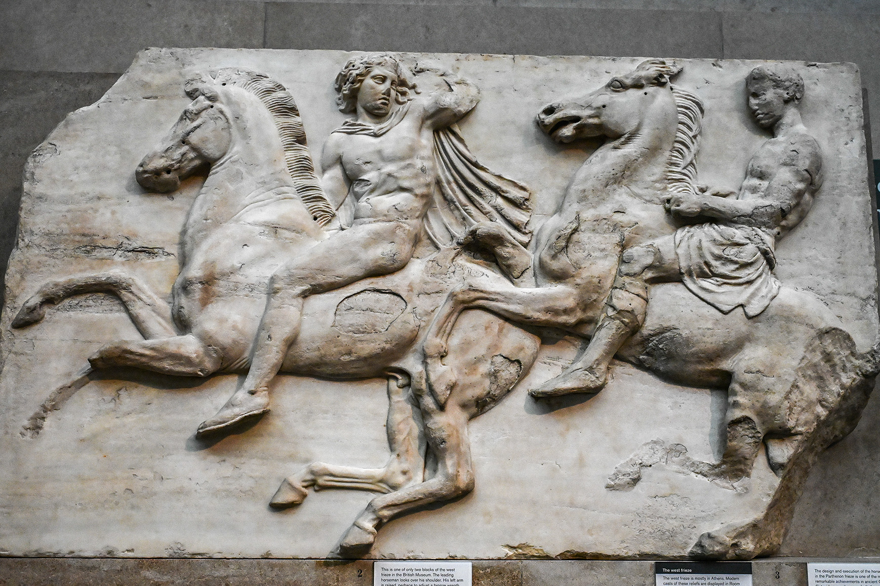 The image depicts a marble relief sculpture featuring two men riding horses. The central figure is a bearded man, likely a warrior, holding a spear and a shield, riding a horse. He is accompanied by another figure, possibly a younger man or a boy, who is also on horseback. The sculpture is part of the west frieze of the Parthenon and is currently housed in the British Museum. The relief showcases the detailed craftsmanship and artistic style characteristic of ancient Greek art, highlighting the dynamic movement and anatomical precision of the figures.