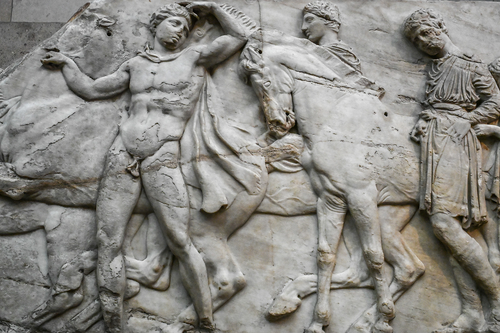 The image depicts a classical Greek sculpture, likely a marble relief, featuring three human figures and a horse. The central figure is a muscular man holding a horse by its reins, while the other two figures, dressed in draped garments, appear to be engaged in some form of activity or interaction. The art style and attire suggest it is from ancient Greece, possibly representing a mythological or historical scene.