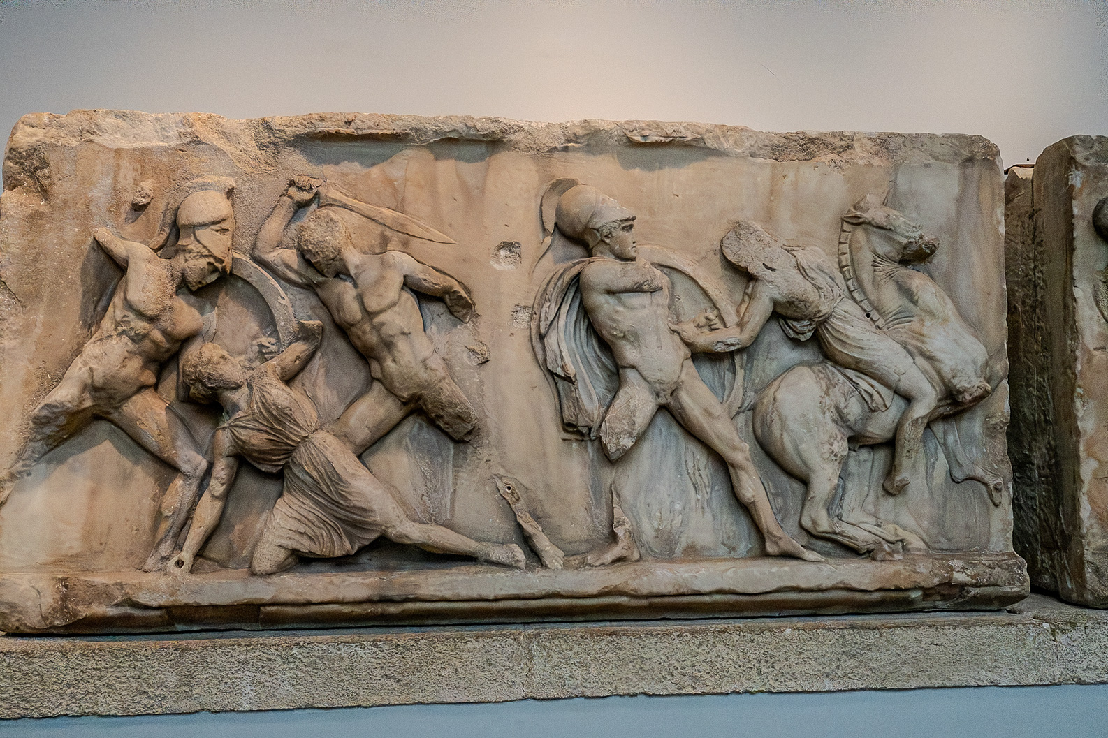 The image depicts an ancient Greek relief sculpture showing a battle scene with various figures engaged in combat, including soldiers and a horse. The sculpture is intricately detailed, capturing the dynamic movement and intensity of the battle. The figures are shown in a combination of defensive and offensive poses, highlighting the chaos and action of the scene. The relief is likely part of a larger architectural element, possibly from a temple or monument, and showcases the artistic skill of the period.