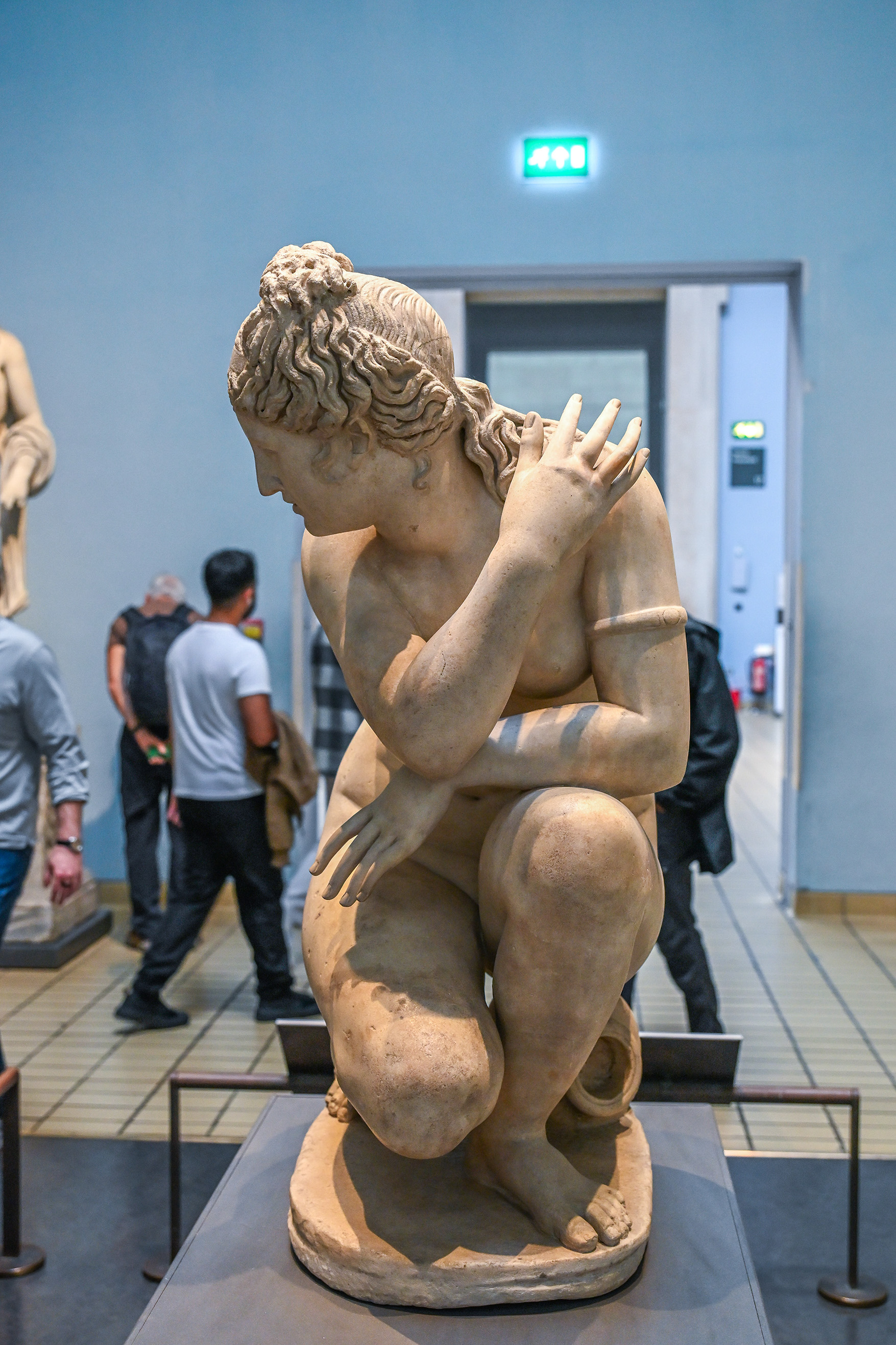 The image shows a classical statue of a seated figure, likely from ancient Greek or Roman art, displayed in a museum. The statue depicts a muscular male figure in a contemplative pose, with one arm resting on its knee and the other hand supporting its head. The figure has detailed curly hair and is shown in a dynamic, slightly twisted position. The statue is placed on a pedestal and is cordoned off with ropes to prevent visitors from touching it. In the background, several people are observing the statue and other artworks in the museum.