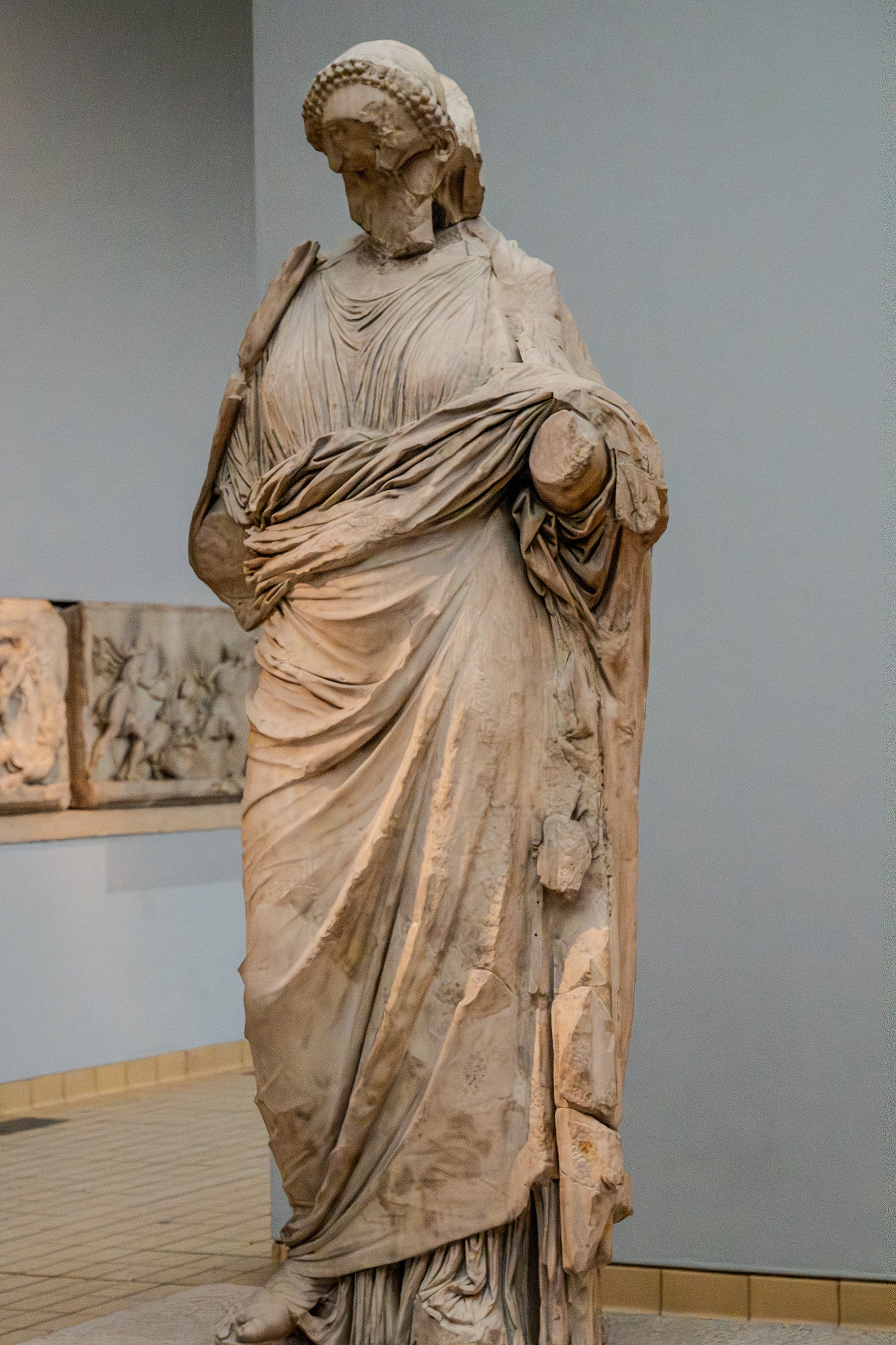 The image depicts a classical statue of a robed figure, likely from ancient Greece or Rome, displayed in a museum. The statue is made of marble and shows a person draped in elaborate, detailed clothing. The figure's head is adorned with a braided hairstyle, and the statue appears to be in a standing pose with one arm bent at the elbow. The background includes a wall with a relief sculpture and a tiled floor.