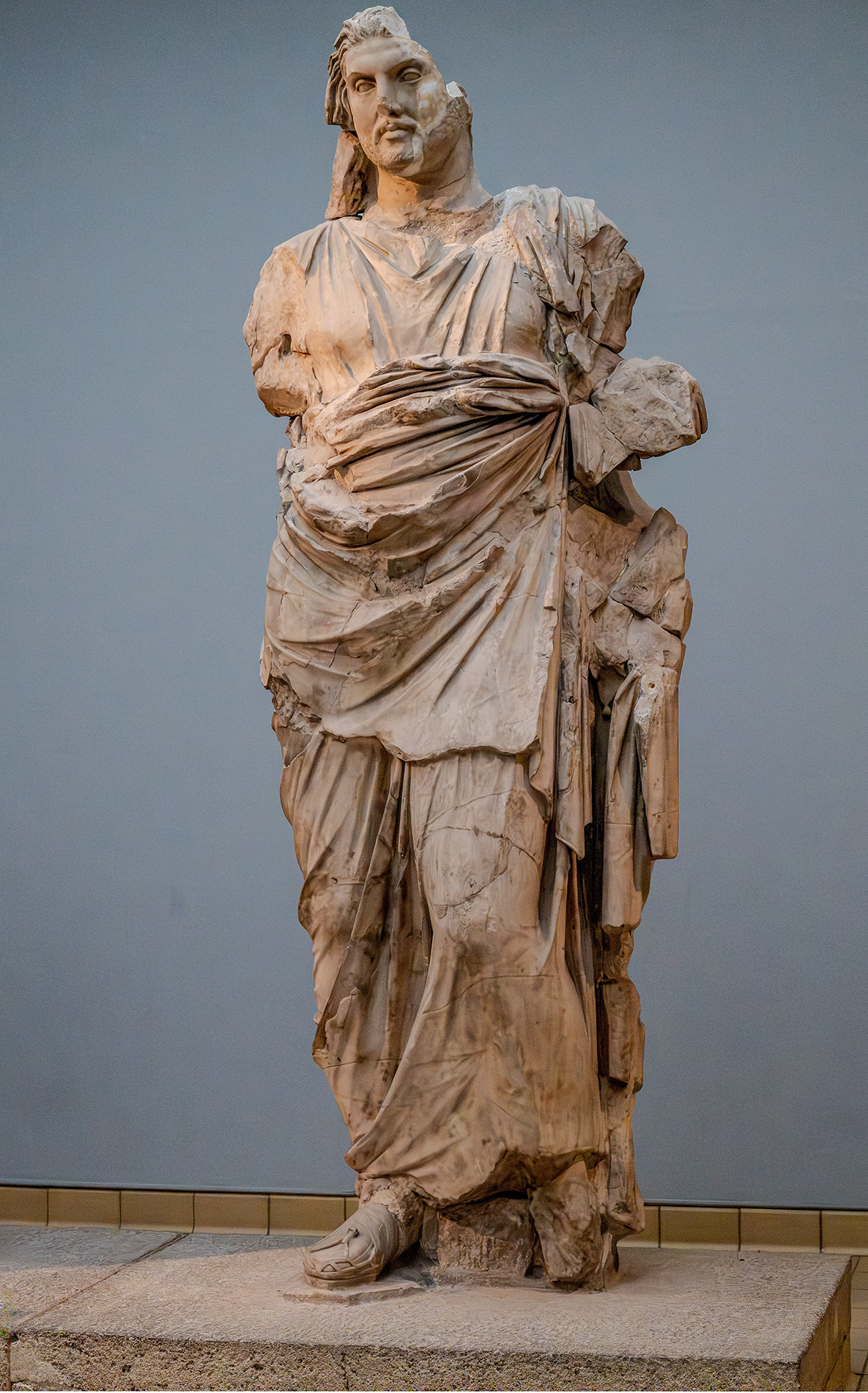 The image depicts a large, ancient statue of a robed figure, possibly from classical antiquity. The statue is made of stone and shows significant wear and damage, with parts of the figure's limbs and drapery missing or broken. The figure is standing upright with one arm bent at the elbow, holding what appears to be a scroll or a similar object. The statue is displayed indoors, likely in a museum setting, with a plain background and a tiled floor.