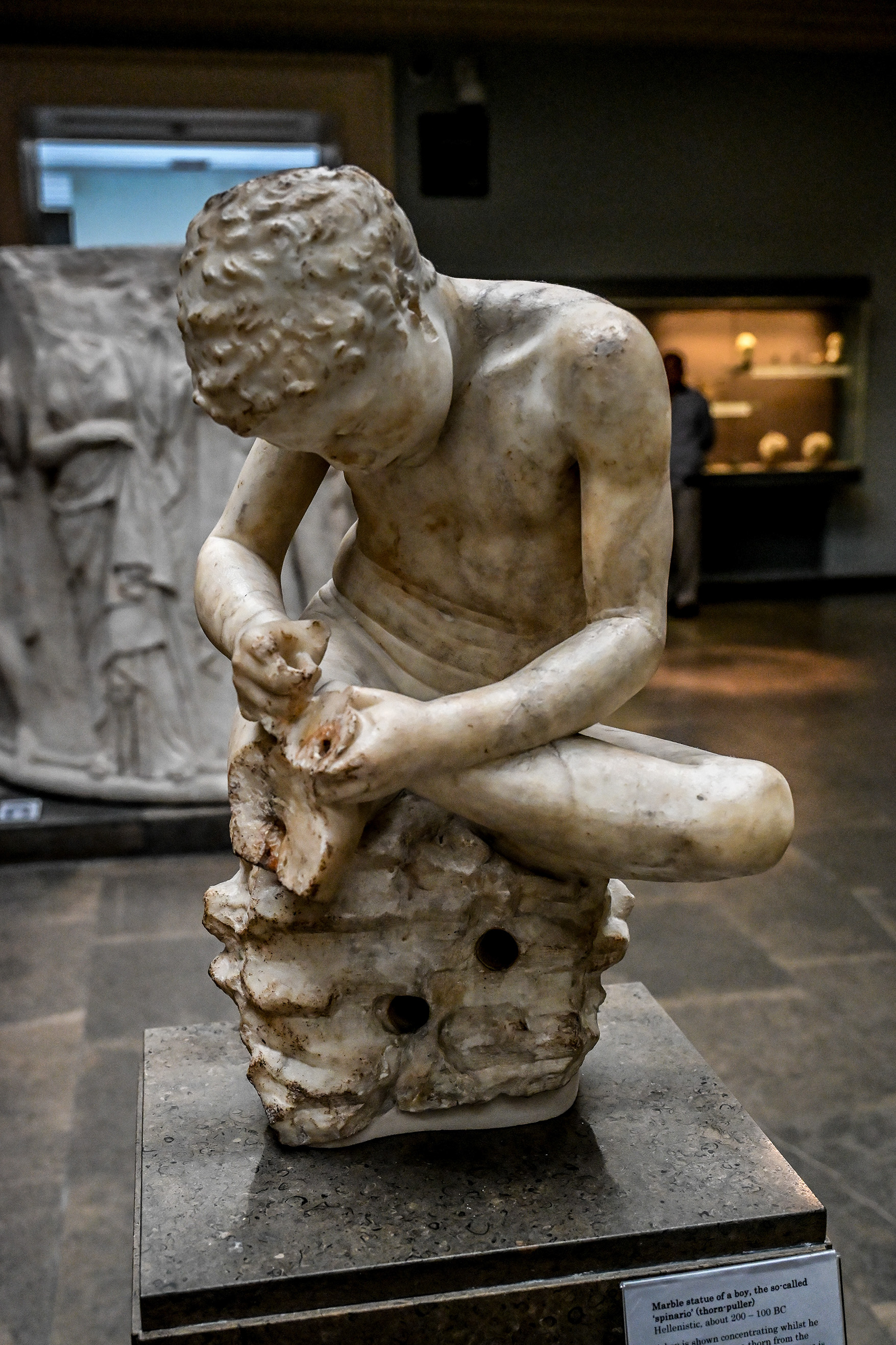 The image shows a marble statue of a boy, known as the 'spinario' or thorn puller, from the Hellenistic period around 200-100 BC. The statue depicts the boy in a concentrated pose as he removes a thorn from the sole of his foot. The statue is displayed in a museum setting, with a descriptive plaque in front of it.