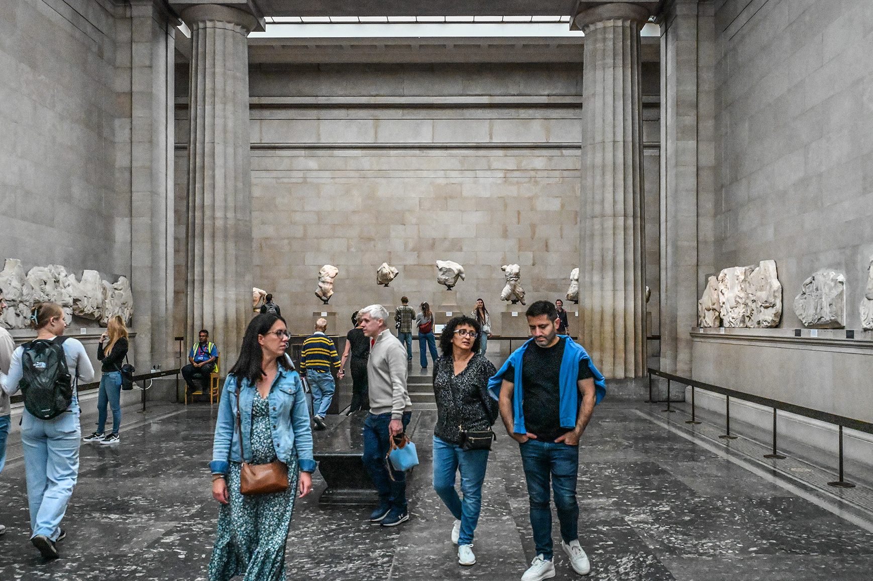 The image depicts a group of people walking through a museum gallery adorned with ancient stone carvings and sculptures. The gallery features large stone columns and fragments of classical sculptures mounted on the walls. Visitors are seen observing the exhibits, with some standing and others walking through the space. The atmosphere suggests a quiet, contemplative environment typical of a museum setting.