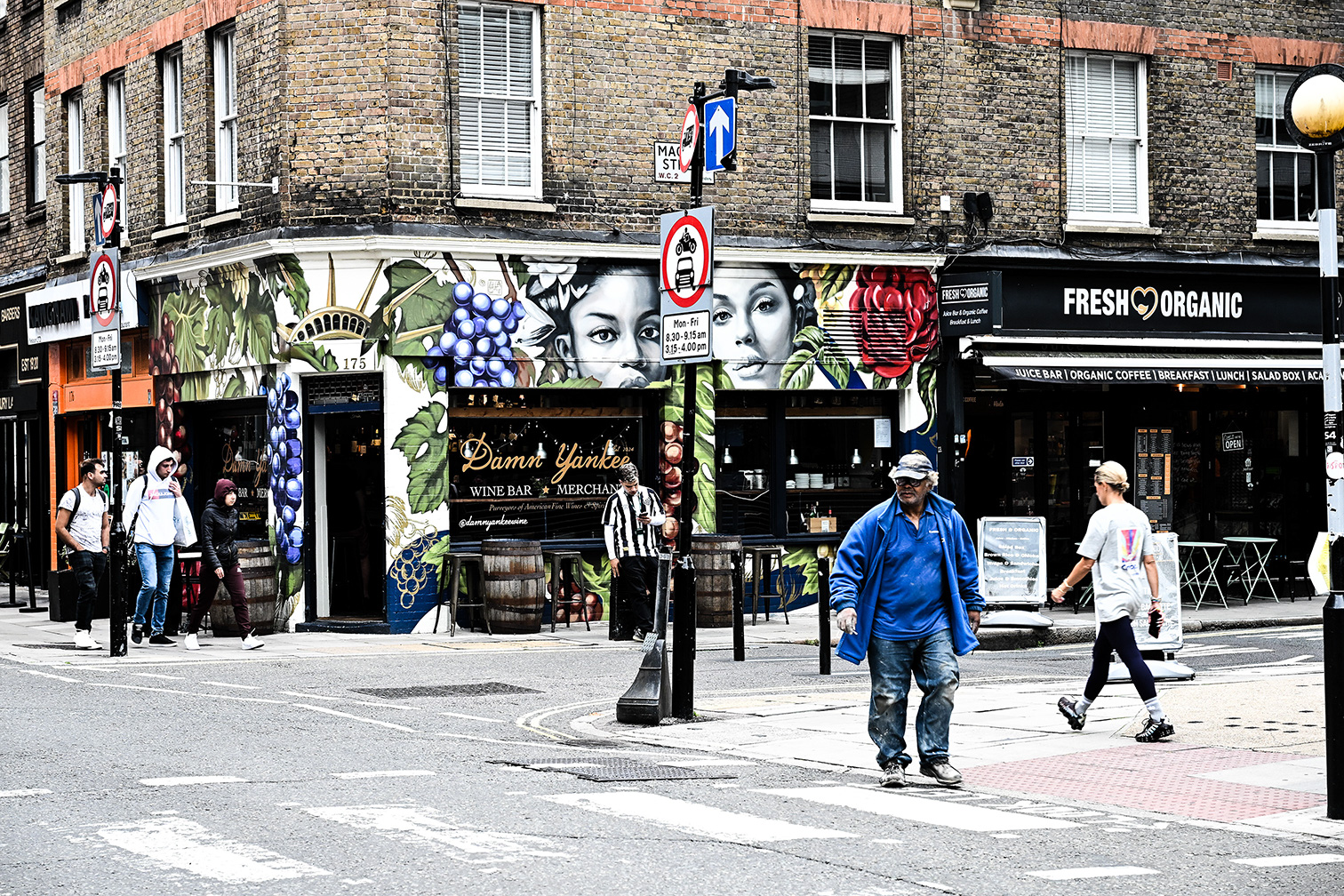 The image depicts a street scene with several pedestrians and storefronts. Prominent businesses include 'Damn Yankee,' a wine bar and merchant, and 'Fresh Organic,' offering organic food and beverages. The street is bustling with activity, and various signs and decorations adorn the buildings, contributing to a vibrant urban atmosphere.