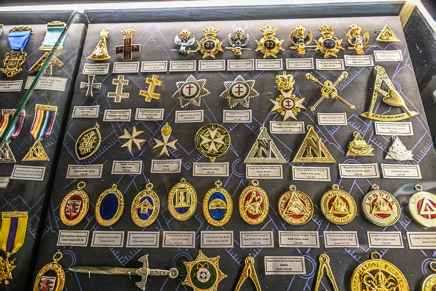 The image shows a display of various Masonic regalia and jewelry, including medals, collars, and other insignia. Each item is labeled with its name and price. The items are arranged neatly in rows and columns, showcasing a variety of designs and symbols associated with Freemasonry.