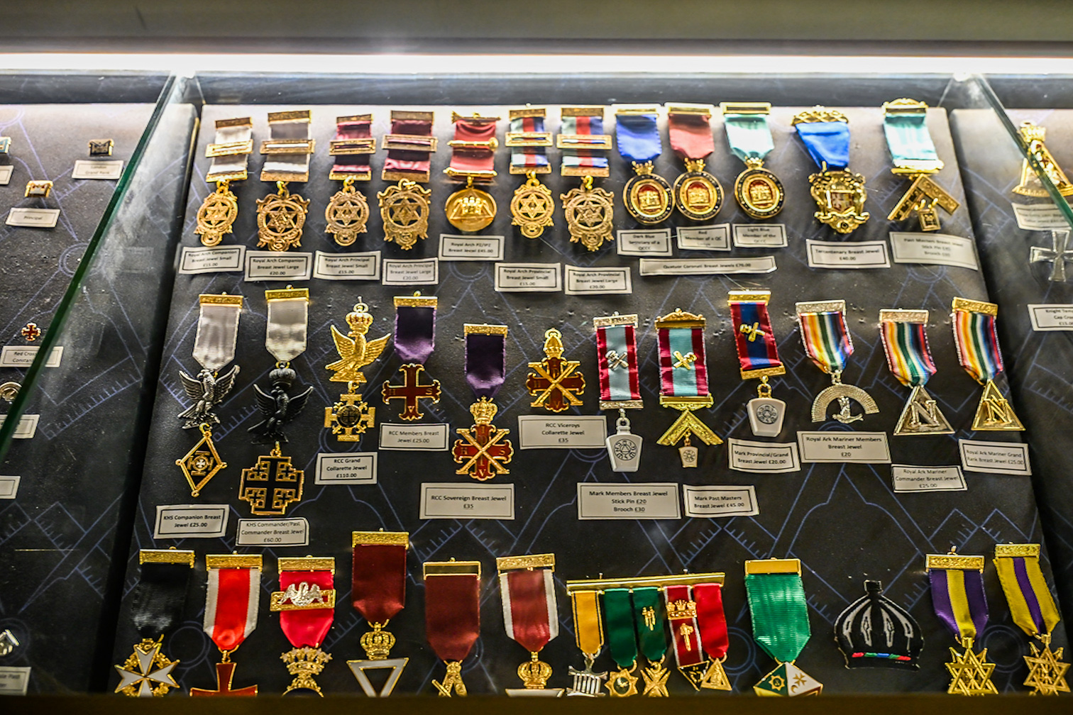 The image displays a collection of various medals and insignia, each meticulously arranged and labeled with descriptions and prices. The medals vary in design, color, and size, showcasing intricate details and craftsmanship. They are organized in rows, with each medal having a small placard in front of it providing information about the item. The display appears to be part of a museum or exhibition, highlighting the historical and cultural significance of these medals.