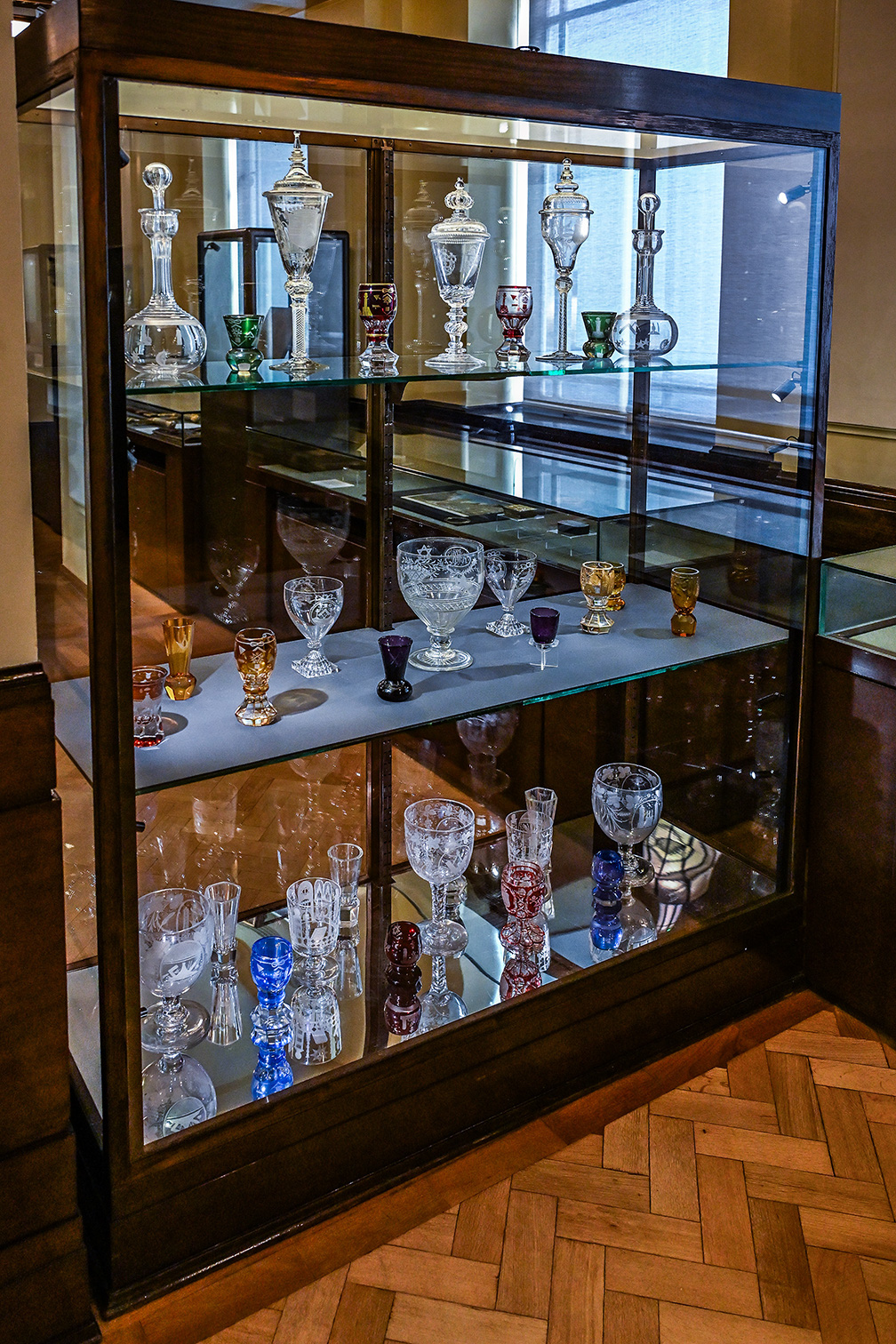 The image shows a glass display case containing a collection of various glassware items. The case has three shelves, each holding different types of glasses, goblets, and decorative pieces. The items vary in shape, size, and color, including clear, colored, and intricately designed pieces. The display is well-lit, highlighting the craftsmanship and details of each item. The floor beneath the display case is made of polished wood, adding to the elegant presentation of the collection.