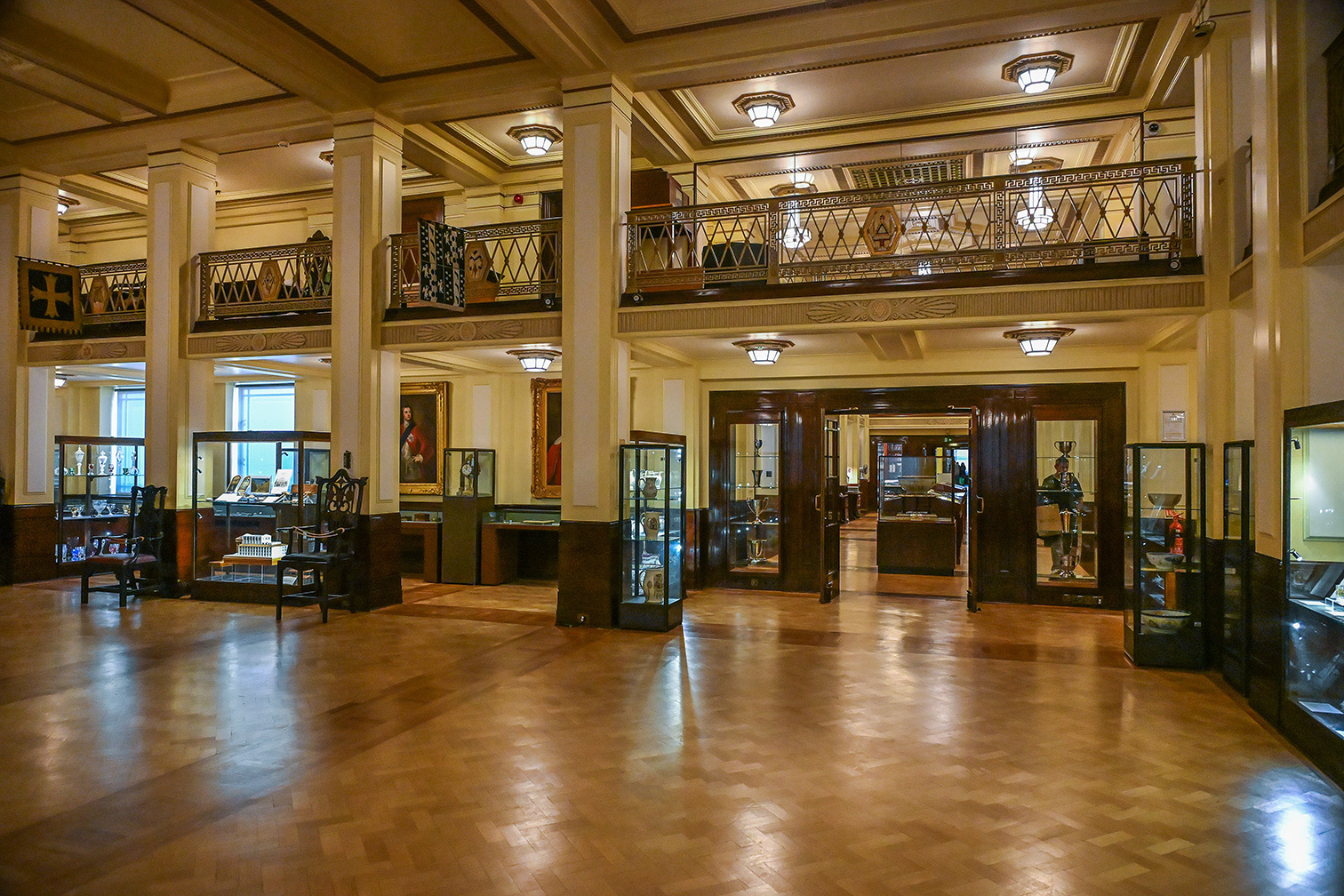 The image depicts an elegant, spacious interior of what appears to be a museum or gallery. The room features high ceilings with ornate designs, large columns, and intricate railings on the upper level. The floor is made of polished wood with a geometric pattern. Display cases filled with various artifacts and pieces of furniture are arranged around the room. The lighting is warm and inviting, with several hanging lanterns illuminating the space.