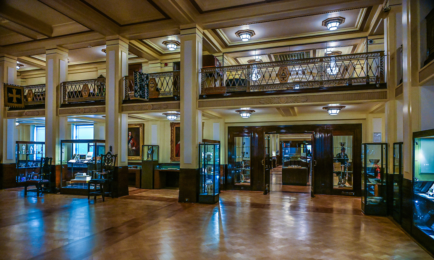 The image depicts an elegant, spacious interior with high ceilings and intricate architectural details. The room features large wooden columns, ornate railings on a balcony, and multiple display cases showcasing various artifacts and items. The floor is made of polished wood, reflecting the light from the lantern-style ceiling fixtures. There are several pieces of antique furniture and artwork on display, including paintings and decorative items. The overall ambiance suggests a museum or a historical building.