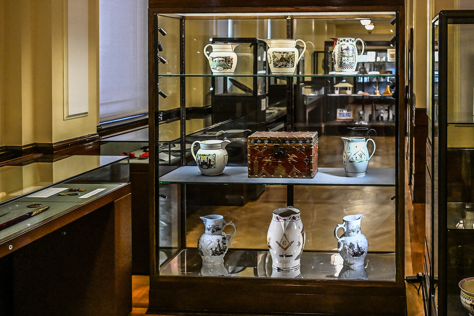 The image shows a display case in what appears to be a museum or an exhibit. The case contains various pieces of pottery, including jugs and a decorative box, all placed on glass shelves. The items are intricately designed and appear to be historical artifacts. The background suggests a well-lit, organized space with other display areas and visitors.