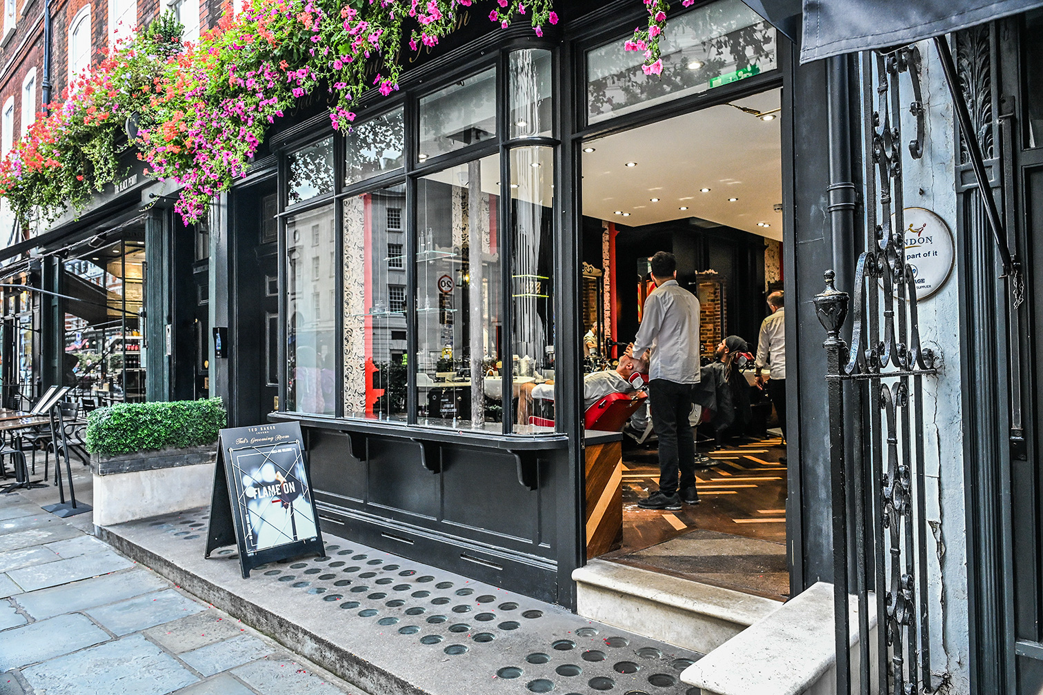 The image depicts the exterior of a restaurant named 'Flamsteed'. The facade features large glass windows with black frames, adorned with hanging flower baskets. The entrance is open, revealing a well-lit interior where people are seated and standing. A sandwich board outside advertises '1/2 Price Cocktails'. The setting appears to be urban, with a sidewalk and street visible in the foreground.