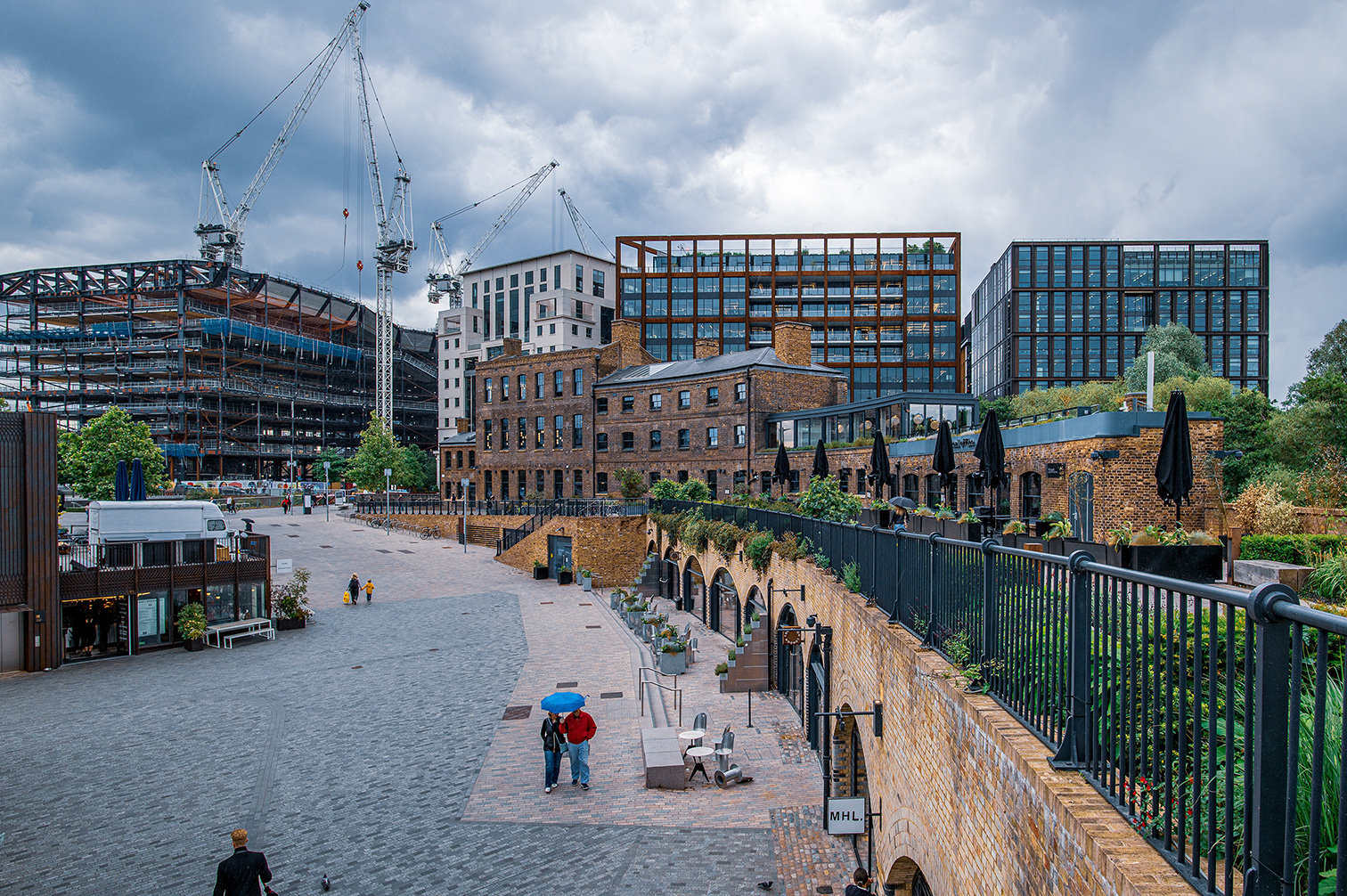 The image depicts a modern urban area under development with a mix of old and new architecture. Construction cranes are visible in the background, indicating ongoing building work. The scene includes a pedestrian walkway with a couple holding an umbrella, surrounded by greenery and outdoor seating areas. The overall atmosphere suggests a blend of historical and contemporary elements in a cityscape.