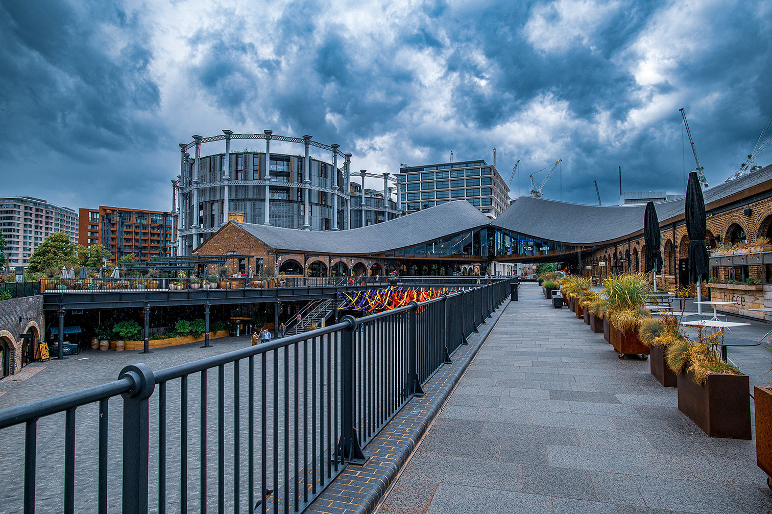 The image depicts a modern urban scene featuring a pedestrian walkway lined with railings and large potted plants. The walkway leads to a large, architecturally distinct building with a combination of brick and glass elements. In the background, there are several high-rise buildings and cranes, indicating ongoing construction. The sky is overcast with dark, heavy clouds, suggesting impending rain. The area appears to be a mix of commercial and public spaces, with outdoor seating and dining areas visible.